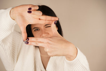 Happy dark haired woman got inspiration to imagine hot to take an interesting picture. Taking a picture with a gesture, smiling with pleasure. Dressed in casual clothes, isolated on beige background.の写真素材