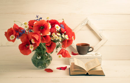 Red poppies bouquet in round vase, open book, tea cup and photo frame on background of white wooden planks in scandinavian styleの写真素材