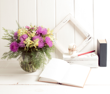 Flowers bouquet in glass round vase with open diary, book, frame and candle on casket, fountain pen on background of white wooden planks in provence style. Light home interior.の写真素材