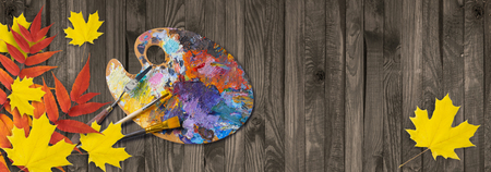 Palette for oil paints with brushes and palette knife on shabby wooden table with autumn leavesの写真素材