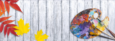 Palette for oil paints with brushes and palette knife on shabby wooden table with autumn leavesの写真素材