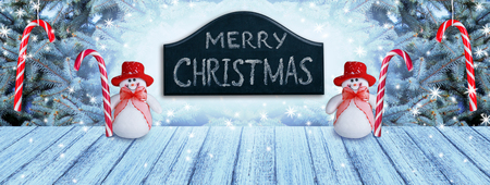 Christmas background with fir tree and two snowmen with candy canes,  wooden table, and snowfall. New Year winter holidays theme. Empty space for your decoration, text or advertising.の写真素材
