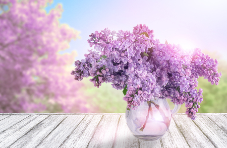 Wooden table in Shabby Chic style with lilac flowers in glass vase and spring trees  against the blue skyの写真素材