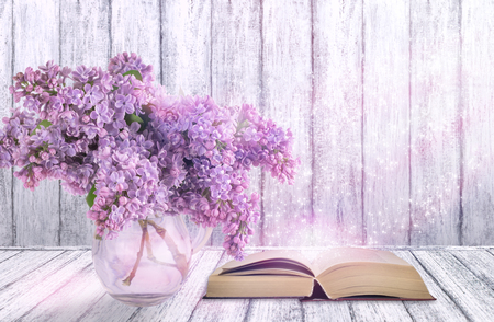 Room interior with lilac flowers in glass vase and open fairytale book on table in shabby chic style on wooden wall background with place for your decoration or text. の写真素材