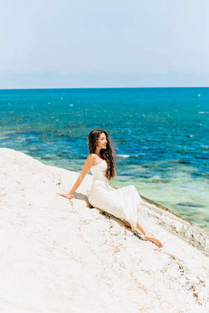 Charming girl in wedding dress standing on stone on sea backgroundの写真素材