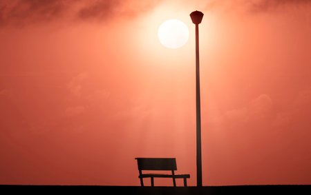 A Wooden bench and street lamp with a background view of a red sunset on sunset and a bright sun .の写真素材