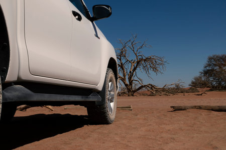 A white off-road car stands in the red sands of the Namib Desert on a sunny day.  Sossusvlei,...