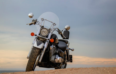 A lone motorcycle stands on the sandy surface against a dramatic cloudy sky. The sunset lighting highlights the contrast between nature and machine.の写真素材