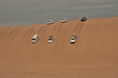 Group of 4x4 off-road vehicles descending steep sand dunes in the Namib Desert, Namibia. Adventure travel in bright sunlight, golden sand, and vast African landscape.の写真素材