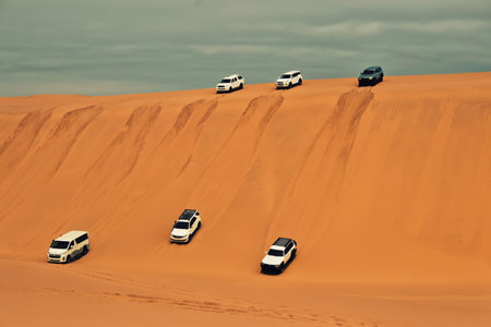 Group of 4x4 off-road vehicles descending steep sand dunes in the Namib Desert, Namibia. Adventure travel in bright sunlight, golden sand, and vast African landscape.の写真素材