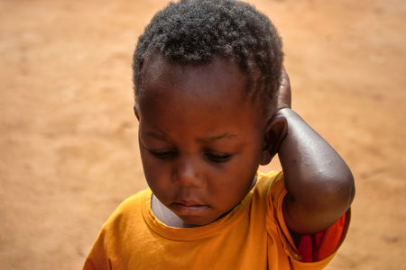 A young Black child  alone in a Namibian refugee camp, his face marked by sadness and quiet...
