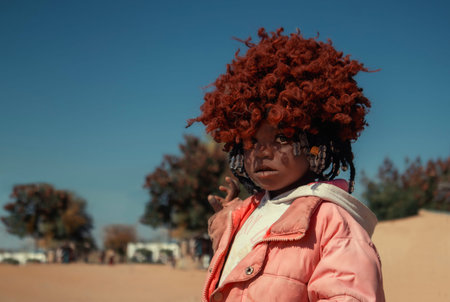 Young African girl wearing a vibrant orange wig and a pink jacket, captured in natural light. A...