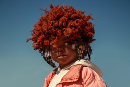 Young African girl wearing a vibrant orange wig and a pink jacket, captured in natural light. A...