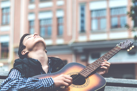 Young man while sitting on the bench playing on acoustic guitar outdoor