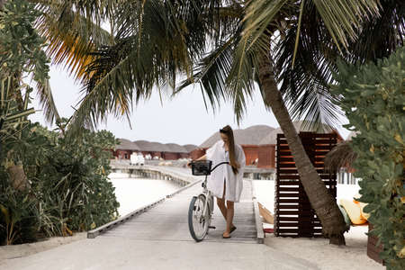 Young girl in white shirt with a bike on the wooden bridge in the Maldives. Bungallow at the background. Island.の写真素材
