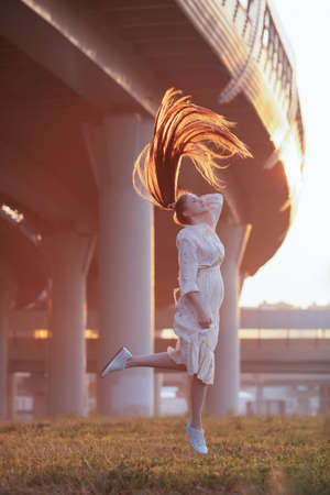 Attractive girl with red long hair, slender and beautiful posing for a photo in a city landscape. Sunrise portrait. Dawn.の写真素材