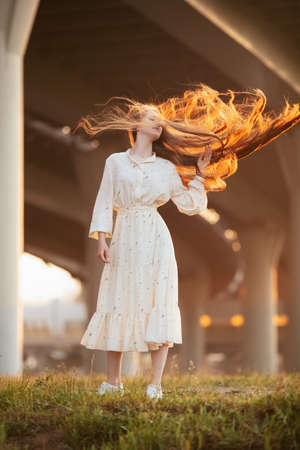 Attractive girl with red long hair, slender and beautiful posing for a photo in a city landscape. Sunrise portrait. Dawn.の写真素材