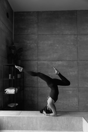 Young sporty attractive woman practicing yoga, doing headstand exercise, variation of salamba sirsasana pose, working out, wearing sportswear, grey top, indoor close up view, yoga studio, rear viewの写真素材