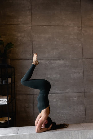 Young sporty attractive woman practicing yoga, doing headstand exercise, variation of salamba sirsasana pose, working out, wearing sportswear, grey top, indoor close up view, yoga studio, rear viewの写真素材