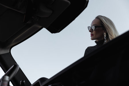 A beautiful girl with blonde hair sits on the hood of a large black car in a dress with an open back. Long hair and black glasses. Black leather jacket in the foreground. Attractive personalityの写真素材