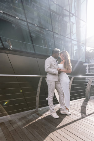 International wedding. The handsome dark-skinned groom in a white suit embraces a beautiful Caucasian bride with a gorgeous hairstyle against the backdrop of Singapore cityの写真素材