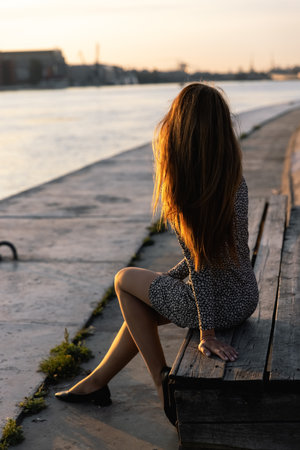 A beautiful girl in a dress with a small print is sitting on a wooden bench on embankment and admiring sunset, girls face is not visible. Soft light highlights her silhouette and beautiful long hair.の写真素材