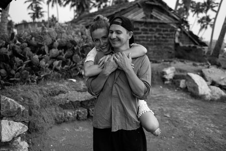 Black and white photo of a young couple embracing on a beach in an island village. The man wears a cap, the smiling woman jumps on his back. Palm trees, cactus, and a fishing hut in the background.の写真素材