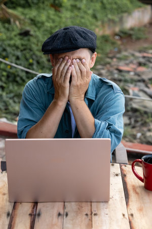 The man covered his face with his hands. A man in a hat is working on a laptopの写真素材