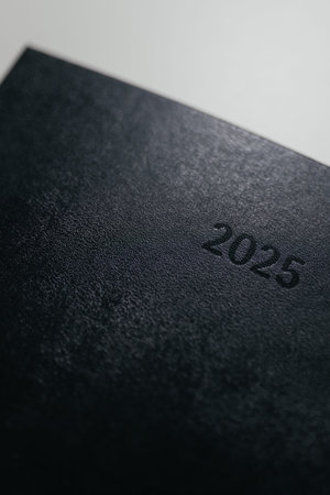 Minimalist black planner or notebook with subtle 2025 embossed text on the cover. Clean, modern...