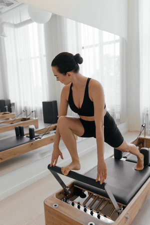 Slim brunette woman in black tight sportswear working out on Pilates reformer machine