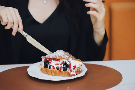 A woman with light skin in a black dress slices a fresh croissant revealing creamy filling and mixed...