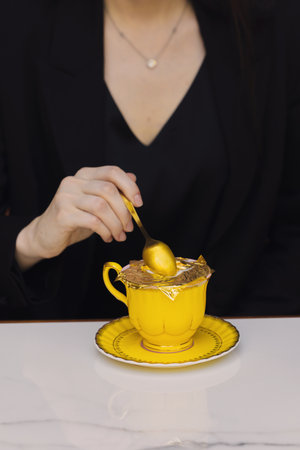 woman in a black blazer stirs golden cappuccino with a small spoon revealing rich golden foam and real edible gold flakes creating an elegant luxurious drink.の写真素材