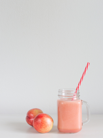A glass jar with beautiful pink smoothie, straw and two peaches on the table. White and pastel pink colors. Minimal design. Copy-space for your text.の写真素材