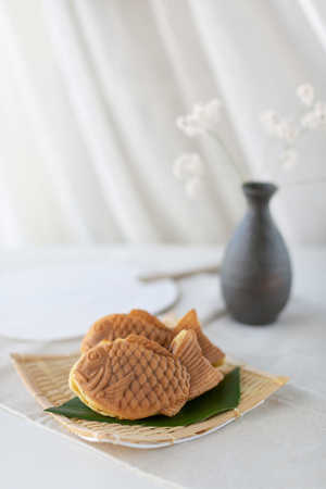 Japanese fish shape cake taiyaki on the traditional bamboo plate. The fillings is sweet red beans azuki. Japanese street food. Decorated with flowers and a fan.の写真素材