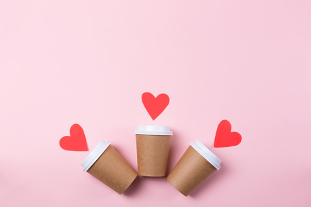 Row of three brown craft cups of coffee or tea to go on the pastel pink background. Flat-lay, top-view. Decorated with red paper hearts. Copy-space.の写真素材