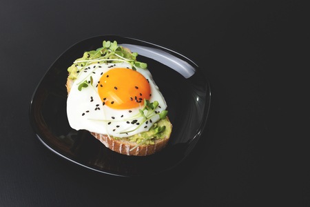 Stylish breakfast with avocado, micro greens and egg toast. Trendy presentation on black plate and gold cutlery. Healthy eating, plant based food concept.の写真素材