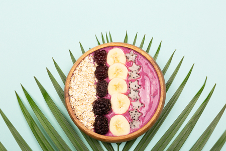 Smoothie or asai bowl on palm leaf. Mint color background. Decorated with berries, bananas, oatmeal, dragon fruit stars, coconut flakes. Healthy breakfast concept. Flat lat, top view.の写真素材