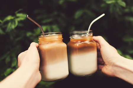 Two glass jars of ice coffee metal straws in woman and man hands. Clink cups of coffee.  Outdoor shot. Lifestyle, picnic vibes. Trendy toning. Copy space for your text.の写真素材