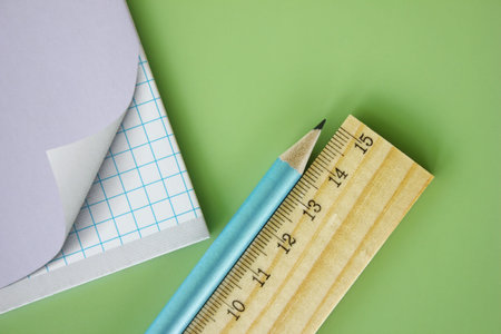 Wooden ruler and pencil are near the school notebook on a green background. Back to school.