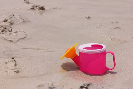 Toy pink watering can with a lid in the shape of a heart is located on the wet sand of the sea coast on a Sunny summer day.の写真素材