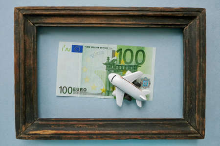 The toy airplane is located on a 100 Euro banknote in a vintage wooden frame on a blue background. The concept of travel. The idea of buying a plane ticket.の写真素材