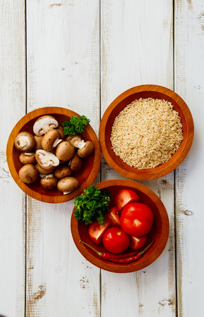 Top view of three wooden bowls with raw rice, mushrooms and tomatoes for cooking over white wooden backgroundの写真素材