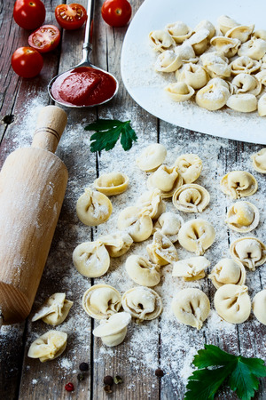 Freshly made italian pasta ravioli on old wooden table with flour, parsley, tomatoes and vintage kitchen accessories.の写真素材