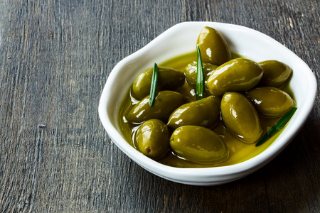whole fresh green olives in a bowl of olive oil on a rustic wooden tabletop, selective focusの写真素材