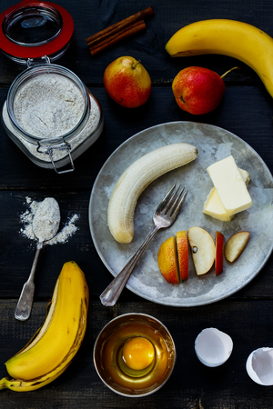 Baking ingredients for cake (flour in glass jar, eggs, butter and fruits)  over rustic wooden board from above. Cooking concept.の写真素材