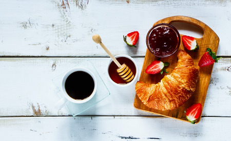 Top view of rural breakfast with cup of coffee, croissant, jam, honey and strawberries on rustic white wooden table. Background layout with free text space.の写真素材