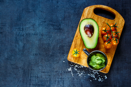 Fresh ingredients for homemade guacamole (avocado, tomato, salt) on rustic wooden cutting board over dark vintage table, top view.の写真素材
