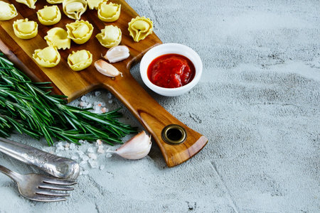 Close up of italian food background with wooden cutting board, homemade raw tortellini, tomato sauce and rosemary over concrete textured board, place for text, border, selective focus.の写真素材