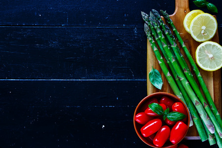 Close up of green asparagus, tomatoes,lemon and basil for tasty asparagus cooking. Green asparagus preparation on dark rustic background, top view, place for text, borderの写真素材
