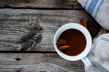 White mug of hot chocolate with cinnamon sticks on rustic wooden background. With space for text,...